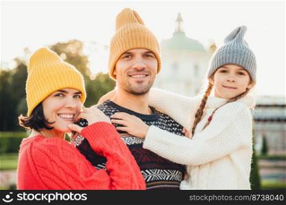 Outdoor portrait of friendly family stand close to each other, have broad smiles. Pretty woman in yellow trendy hat and red sweater, handsome young man holds little adorable daughter on hands
