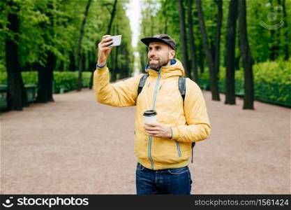 Outdoor portrait of handsome guy with thick beard wearing yellow anorak and jeans holding rucksack, coffee and smartphone making selfie against green trees. Pleased tourist having rest in park