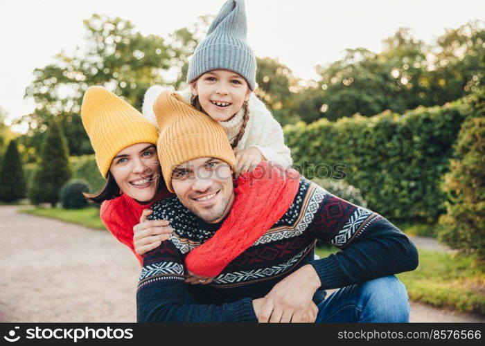Outdoor portrait of handsome man give piggyback to his wife and daughter, wear warm clothes, have happy expressions, support each other. Family embrace outdoors, smile pleasantly into camera