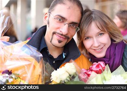 outdoor portrait of man in glasses and beauty blond girl with flower bouquets, looking at camera and smiling
