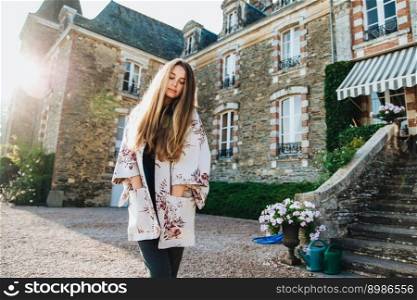 Outdoor portrait of pensive thoughtful beautiful woman with long staight luxurious hair keeps hands in pocket of mantlet, looks down, stands against ancient castle or builduing, wonderful exterior