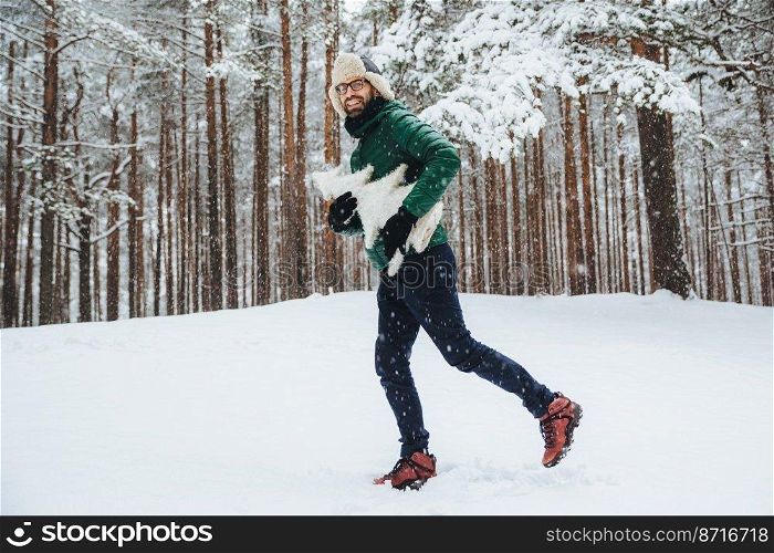 Outdoor portrait of positive happy male has fun, walks in winter forest, enjoys beautiful landscapes and fresh cold weather, holds artificial white firtree, looks directly into camera. Winter concept