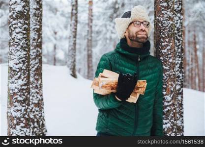 Outdoor portrait of smiling male stands near tree covered with snow, holds firewood, looks happily upwards as notices squirrel on tree, stands in winter forest. People and recreation concept