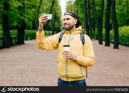 Outdoor portrait of stylish man with stubble beard wearing yellow anorak and holding backpack and takeaway coffee making selfie with his cell phone while standing in beautiful green park having smile