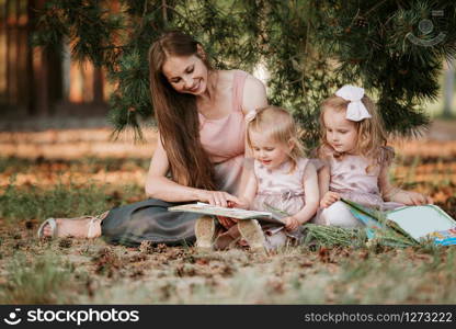 Outdoor portrait of two little girl is reading a book on the grass with mother. She has a look of pleasure and she looked very relaxed in her mother&rsquo;s arms.