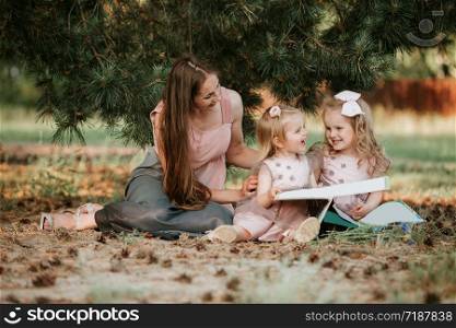 Outdoor portrait of two little girl is reading a book on the grass with mother. She has a look of pleasure and she looked very relaxed in her mother&rsquo;s arms.