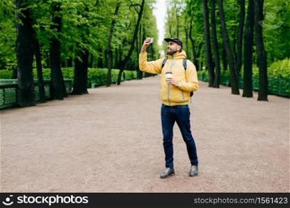 Outdoor portrait of young handsome tourist dressed in stylish clothes holding backpack and takeaway coffee posing into camera of his smartphone making selfie isolated over green nature background