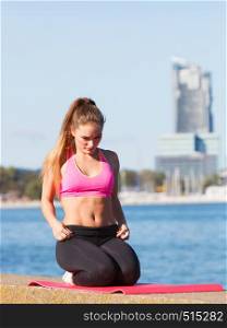 Outdoor relax, sport, fintess concept. Woman in sports suit sitting on dyke, relaxing before workout, water and city in background. Woman in sportswear sitting on dyke by sea