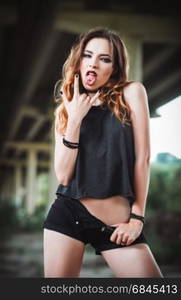 "Outdoor shot: a sexy young rock girl in black shorts and shirt showing tongue and "horns up" gesture"