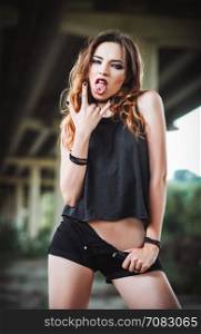 "Outdoor shot: a sexy young rock girl in black shorts and shirt showing tongue and "horns up" gesture"