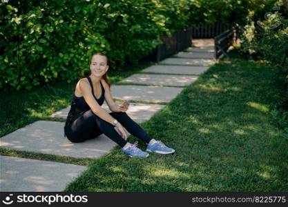 Outdoor shot of active brunette European woman downloads music on smartphone for active training outside has break after workout dressed in active wear shares content in blog looks happily away