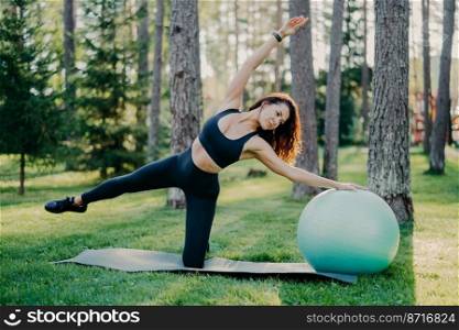 Outdoor shot of active brunette woman in sportswear poses on yoga mat, does stretching exercises with gymnsatic ball, poses in forest or park on green grass. Aerobics, healthy lifestyle concept