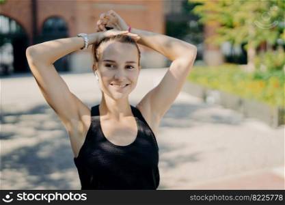 Outdoor shot of active pretty sporty woman combes pony tail smiles gladfully and looks into distance satisfied after cardio training dressed in black tshirt poses outside listens music via earphone