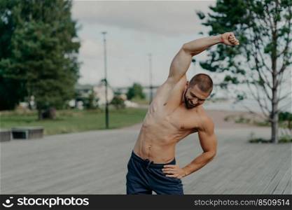 Outdoor shot of activeμscular man makes stretχng excerises and poses with naked torso, trains in open air, has big motivation, keeps fit. Determi≠d sportsman being on way to stron≥r body