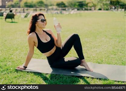 Outdoor shot of active slim woman drinks water from bottle feels thirsty after fitness training concentrated into distance wears cropped top and leggings practices yoga outside. Sport concept