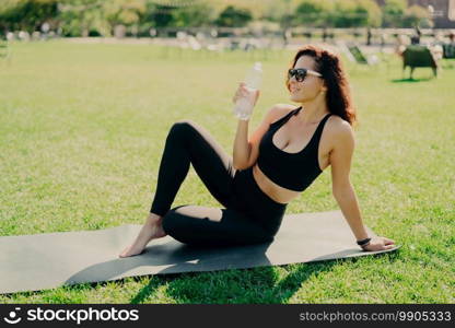 Outdoor shot of active slim woman drinks water from bottle feels thirsty after fitness training concentrated into distance wears cropped top and leggings practices yoga outside. Sport concept