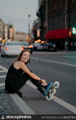 Outdoor shot of active slim woman poses on asphalt puts on rollerskates being in good mood spends free time riding rollerblades in urban place. Blurred city background. Hobby and recreation.