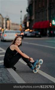 Outdoor shot of active slim woman poses on asphalt puts on rollerskates being in good mood spends free time riding rollerblades in urban place. Blurred city background. Hobby and recreation.