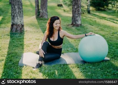 Outdoor shot of active sporty woman takes break after physical exercises, poses on fitness mat with fitball, enjoys fresh air in forest, has perfect body shape uses sport equipment. Gymnastics concept