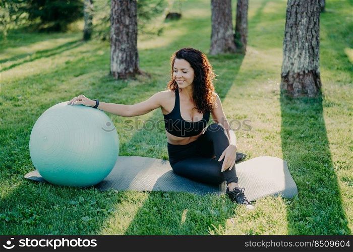 Outdoor shot of active sporty woman takes break after physical exercises, poses on fitness mat with fitball, enjoys fresh air in forest, has perfect body shape uses sport equipment. Gymnastics concept