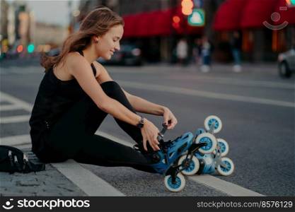 Outdoor shot of active woman laces rollerblades prepares for ride sits on road against busy city background dressed in black activewear enjoys rollerskating. Sporty lifestyle and recreation concept