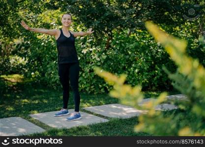 Outdoor shot of athletic fit young woman keeps arms sideways being in good mood exercises outdoors wears sportsclothes enjoys fresh air and beautiful nature. Warming up and exercising concept