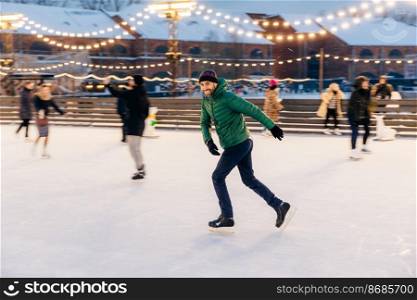 Outdoor shot of attractive man with beard, wears warm winter clothes, practices going skating on ice skate ring decorated with lights, has glad expression, enjoys his favourite action