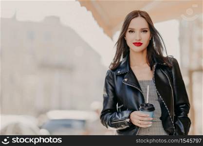Outdoor shot of attractive millennial female model with makeup, dressed in stylish leather jacket, drinks coffee from disposable cup, wanders around city, enjoys leisure time. People, drinking concept