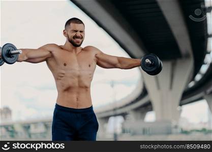 Outdoor shot of bearded European man does biceps curls with barbells, leads healthy lifestyle, clenches teeth as lifts heavy weights, has strong body, poses near bridge. Strong weightlifter.