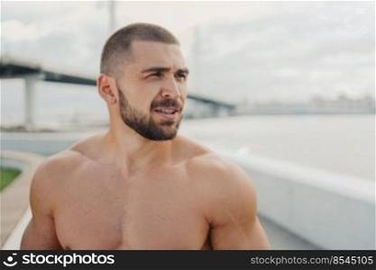 Outdoor shot of bearded man with muscular naked torso concentrated into distance, thinks about gaining sport results, prepares for morning training, has well cared body. Fitness, sport, lifestyle