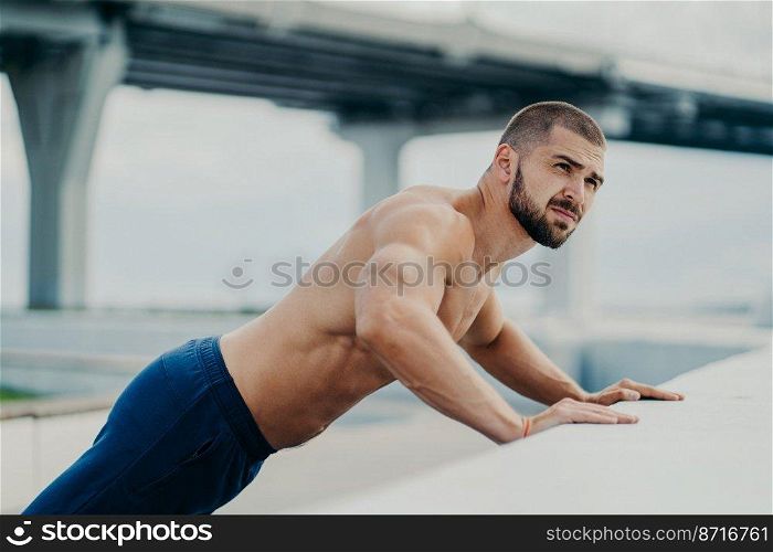 Outdoor shot of bearded muscular man does push up exercises, warms up before training, poses against bridge background, has workout early in morning, concentrated somewhere. Active lifestyle