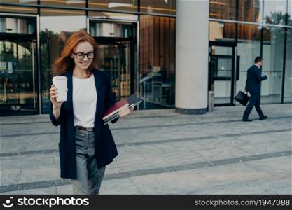 Outdoor shot of beautiful redhead businesswoman carries portable digital tablet notebook for writing notes and disposable cup of coffee walks near office building returns from business meeting. Outdoor shot of businesswoman carries portable digital tablet notebook for writing notes coffee