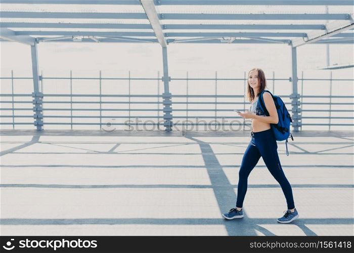 Outdoor shot of beautiful woman wth slim body, dressed in casual top and leggings, carries bag, going in gym, uses modern smart phone for listening music, copy space aside for your advertisement
