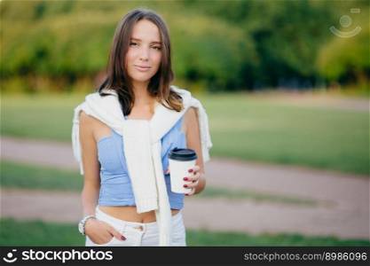 Outdoor shot of brunette woman dressed in casual t shirt, white trousers, keeps hand in pocket, drinks aromatic coffee during morning walk, stands against green nature background. Leisure concept