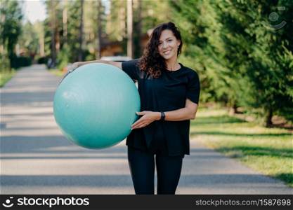 Outdoor shot of brunette woman poses with big fitball, dressed in sportclothes, has training in open air, walks on road, poses near forest. Fitness trainer going to show masterclass for pregnant women