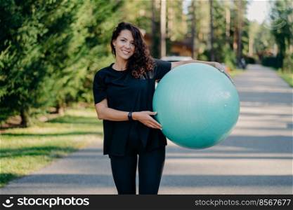 Outdoor shot of brunette woman poses with big fitball, dressed in sportclothes, has training in open air, walks on road, poses near forest. Fitness trainer going to show masterclass for pregnant women