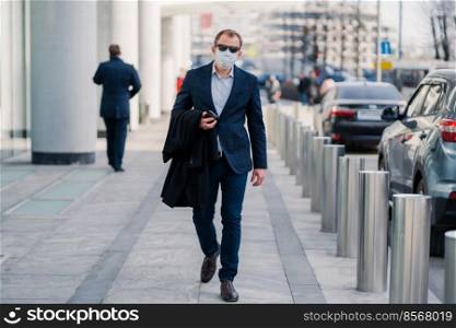Outdoor shot of businessman wears protective medical mask, walks in city street, dressed in formal clothes, holds mobile phone, going to work. Health and safety life from coronavirus and pandemic