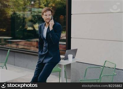 Outdoor shot of busy man entrepreneur solves probems distantly has telephone conversation poses in open air cafe near table with laptop computer and coffee wears formal suit discusses something. Outdoor shot of busy man entrepreneur solves probems distantly has telephone conversation