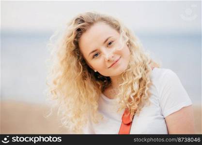 Outdoor shot of curly beautiful young female with fresh skin, looks positively at camera, wears casual white t shirt, poses against blurred seaside background. People and recreation concept.