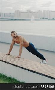 Outdoor shot of determined muscular man warms up before training stands in plank pose has naked torso muscular body enjoys exercising in open air wears active wear has workout to maintain vitality