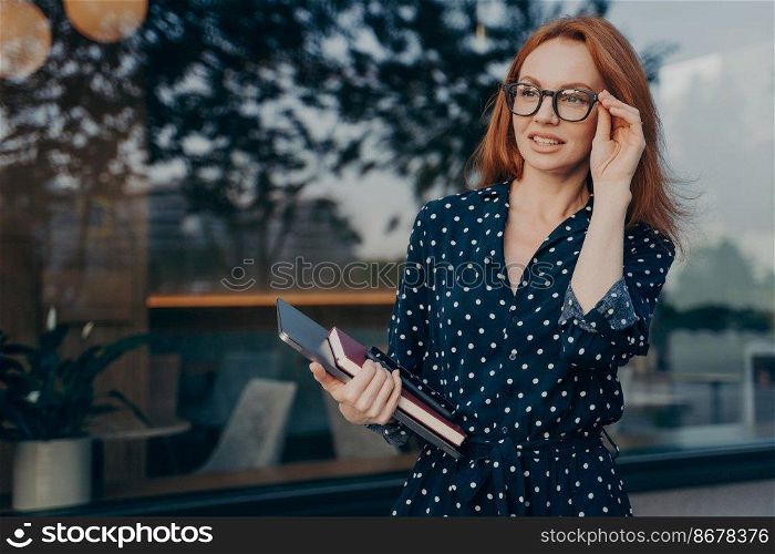 Outdoor shot of female entrepreneur looks attentively into distance keeps hand on spectacles wears black polka dot dress carries tablet smartphone and organizer poses near window of restaurant. Female entrepreneur carries tablet smartphone and organizer poses near window of restaurant