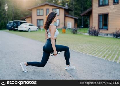 Outdoor shot of fit healthy woman dressed in active wear makes physical exercises with dumbbells in open air trains biceps poses outside with houses in background. Sportswoman enjoys aerobic