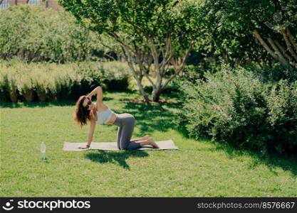 Outdoor shot of fitness woman poses on knees at fitness mat prepares for doing gymnastics dressed in sportswear has regular training outdoors breathes fresh air. Healthy lifestyle wellness exercising