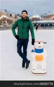Outdoor shot of glad bearded male wears green jacket and hat, stands near skate aid as trying to obtain skating skills, stands on ice rink, has entertainment with friends or family. Active lifestyle