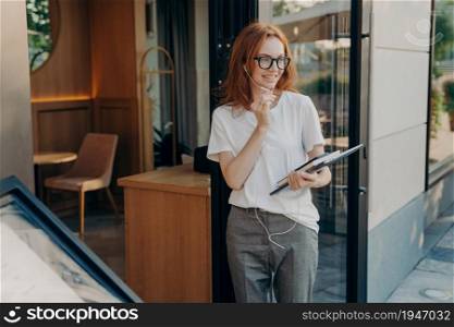 Outdoor shot of glad ginger young woman uses earphones with microphone to communicate holds tablet and notepad satisfied with quality sound in accessory poses in urban place speaks with friend. Outdoor shot of glad ginger young woman uses earphones with microphone to communicate