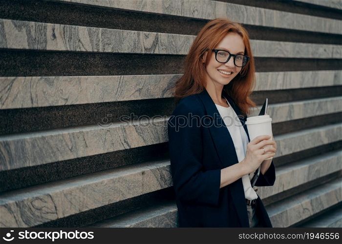 Outdoor shot of glad redhead European woman drinks takeaway coffee holds digital electronic device wears transparent glasses formal clothes stands near marble wall blank space for your text.. European woman drinks takeaway coffee holds digital electronic device stands near marble wall