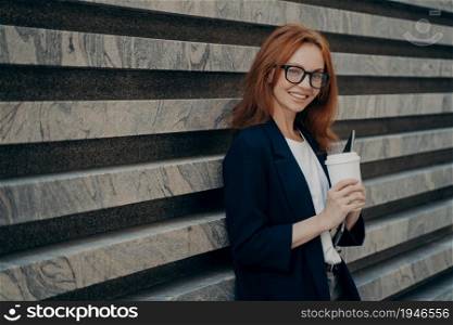 Outdoor shot of glad redhead European woman drinks takeaway coffee holds digital electronic device wears transparent glasses formal clothes stands near marble wall blank space for your text.. European woman drinks takeaway coffee holds digital electronic device stands near marble wall