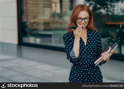 Outdoor shot of glad redhead young woman makes online voice record for sending audio message to business partner holds smartphone near mouth holds notepad and tablet wears polka dotted dress. Woman makes online voice record for sending audio message poses outdoor