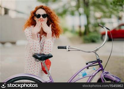 Outdoor shot of gorgeous red haired female covers mouth with both palms, has red manicure, dressed in fashionable dress and trendy sunglasses, poses near her bicycle, spends free time riding bike