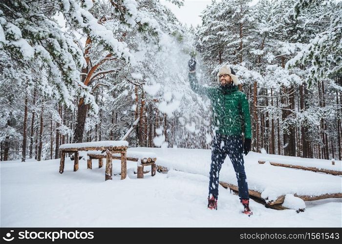 Outdoor shot of handsome bearded male dressed in warm clothes, has fun as throws snow in air, spends holidays in winter forest, expresses positiveness. Recreation and weather concept